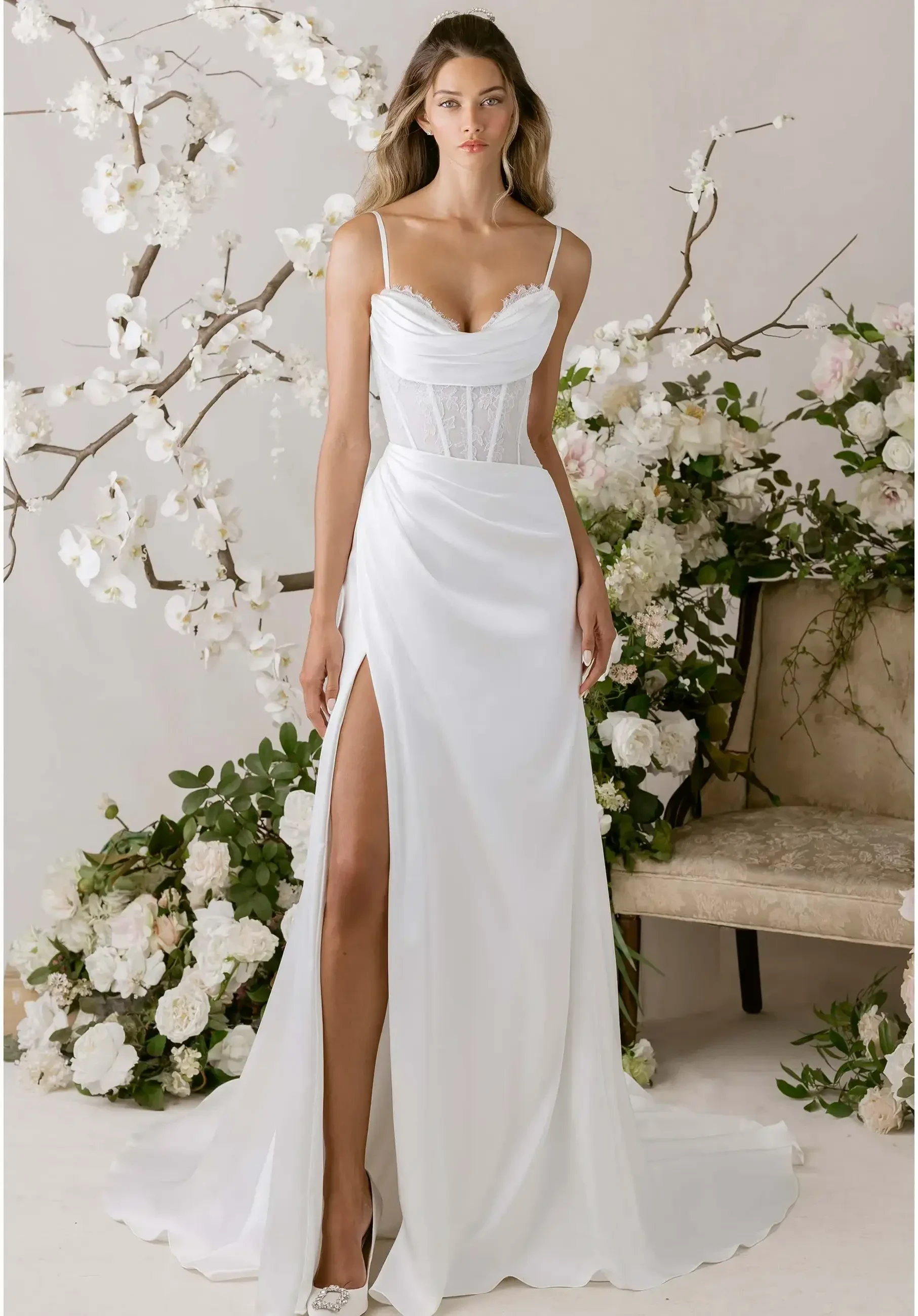 A model wearing a white wedding gown with a thigh-high slit, standing in front of floral decorations and a vintage chair.
