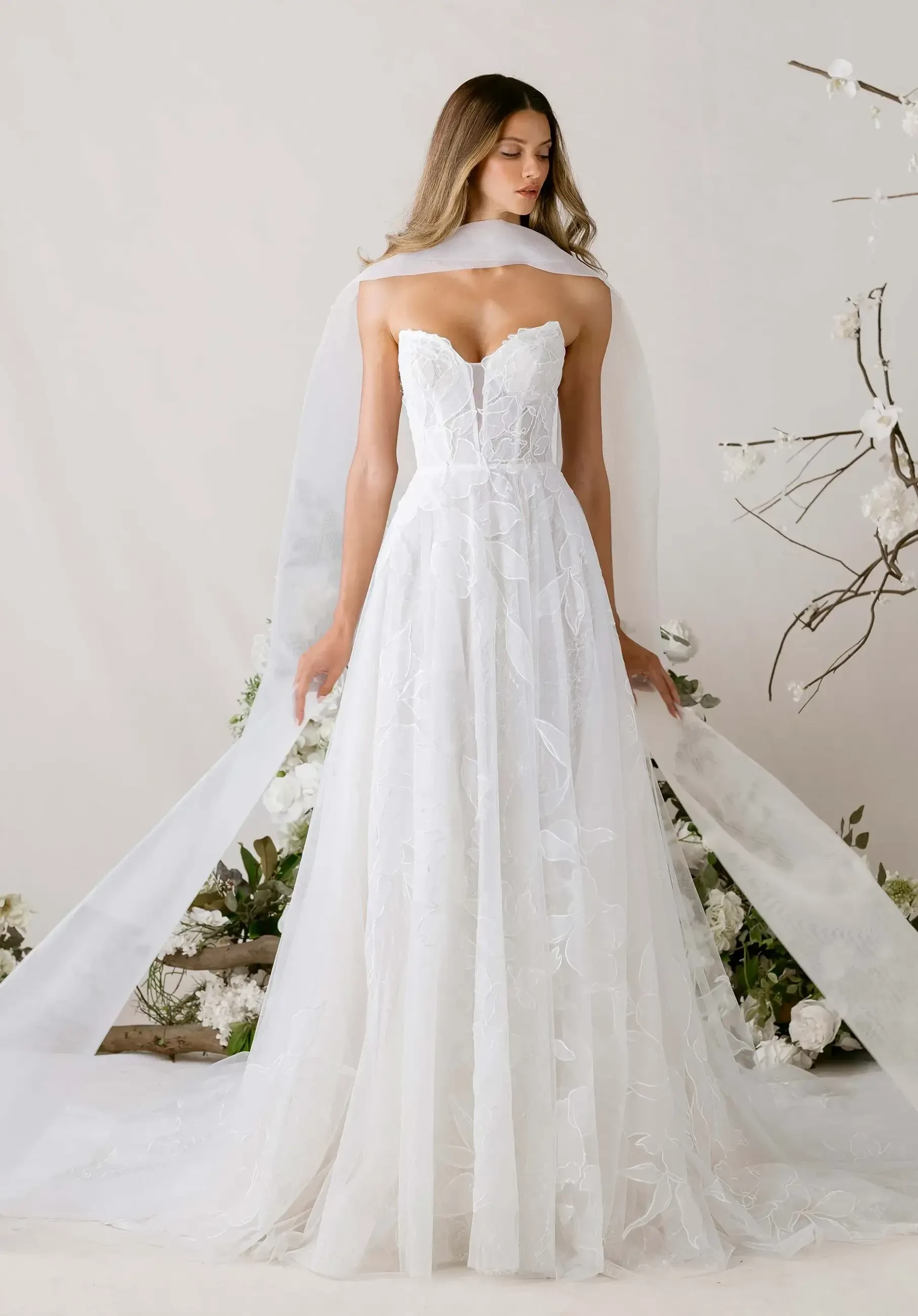 A model wearing a white wedding gown with a strapless bodice, flowing skirt, and a sheer veil, standing against a neutral background adorned with flowers.