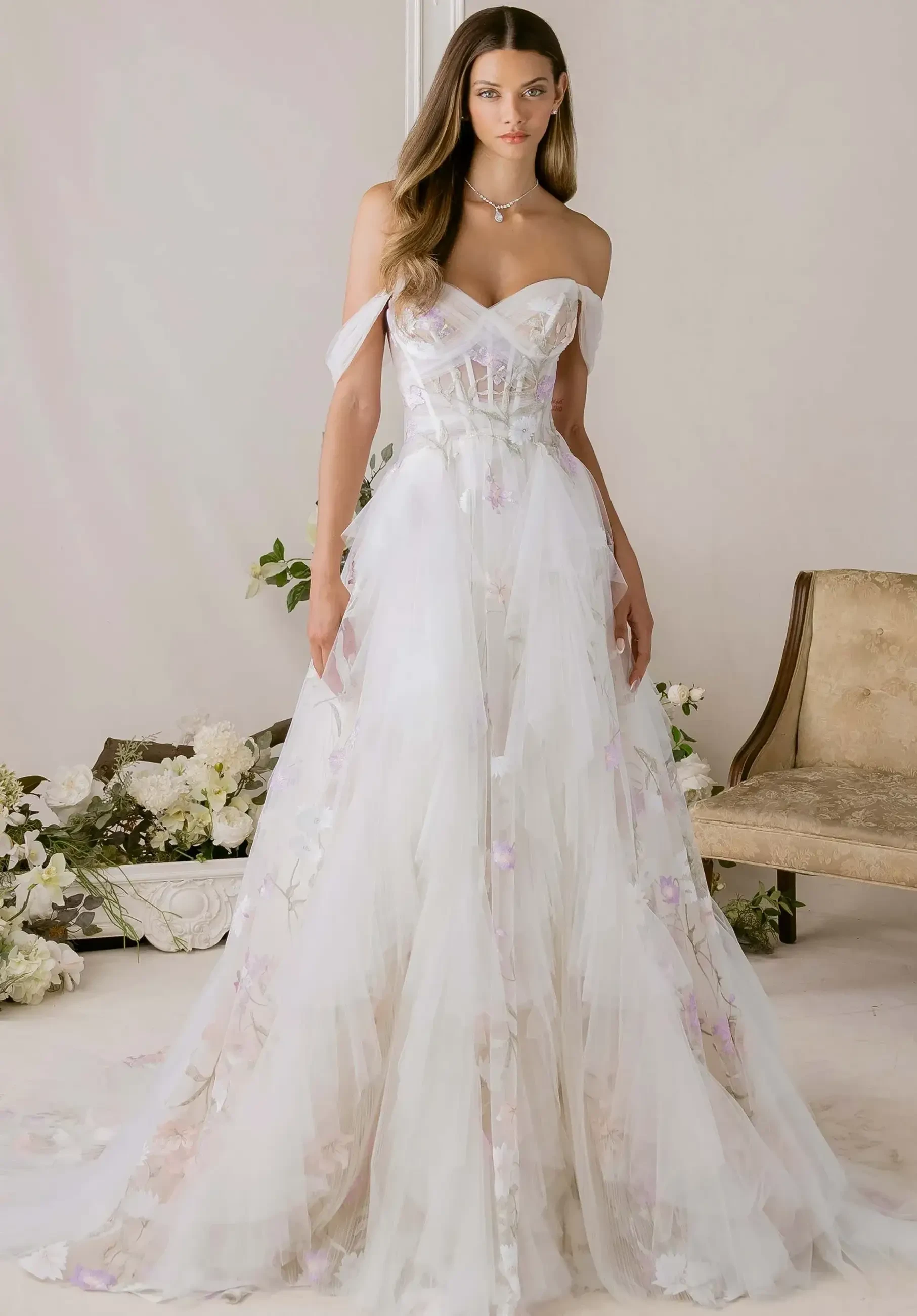 A model is wearing a flowing, off-the-shoulder wedding gown with floral embellishments, standing against a light backdrop.