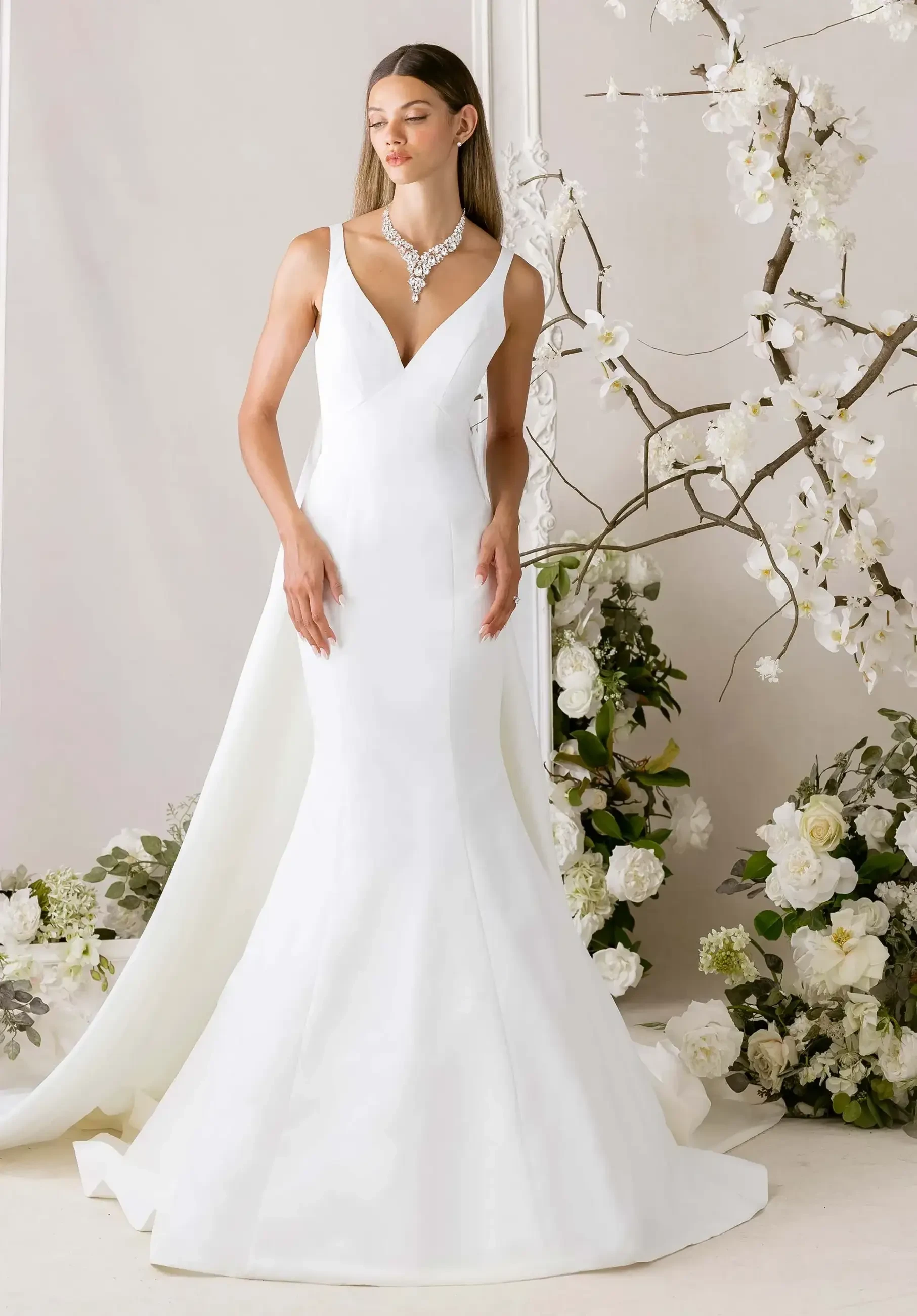 A model wearing a sleek, white wedding gown with a fitted silhouette and a train, accessorized with a statement necklace, standing in front of floral arrangements.