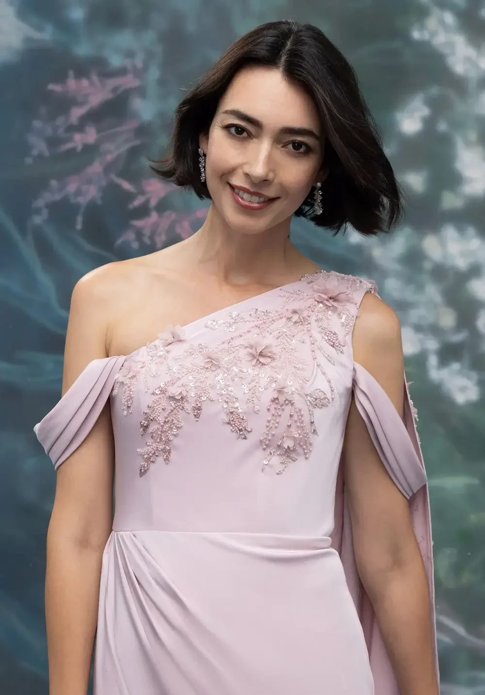 A woman in a light pink evening gown with floral embellishments, standing against a floral backdrop.