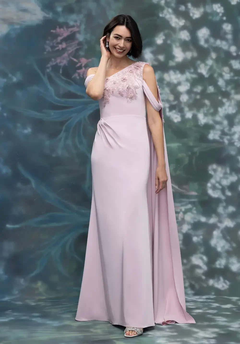 A woman in a light pink evening gown with floral embellishments, standing against a floral backdrop.