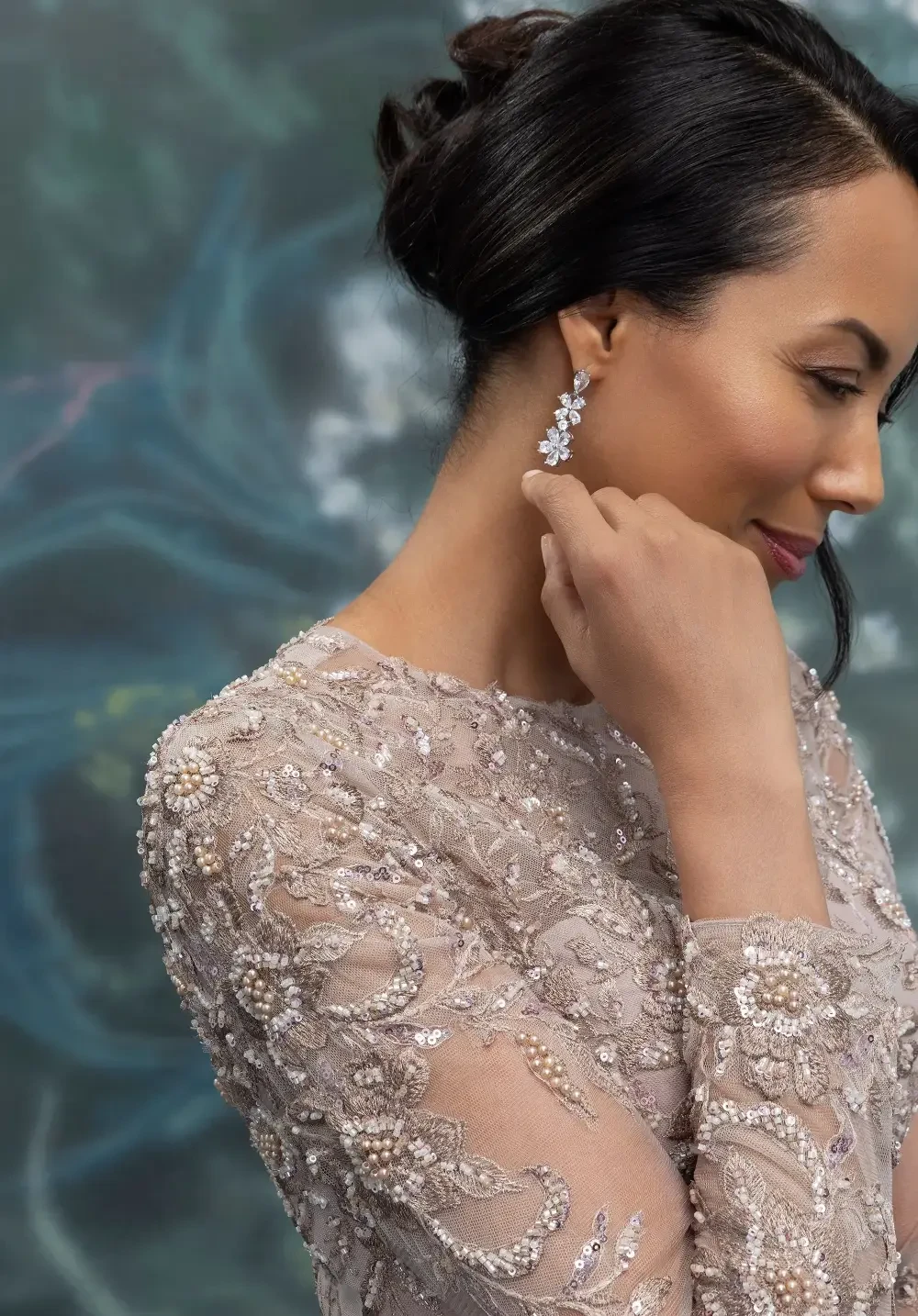 A woman wearing a long, elegant dress with intricate lace detailing on the back, standing against a soft, floral-themed background.