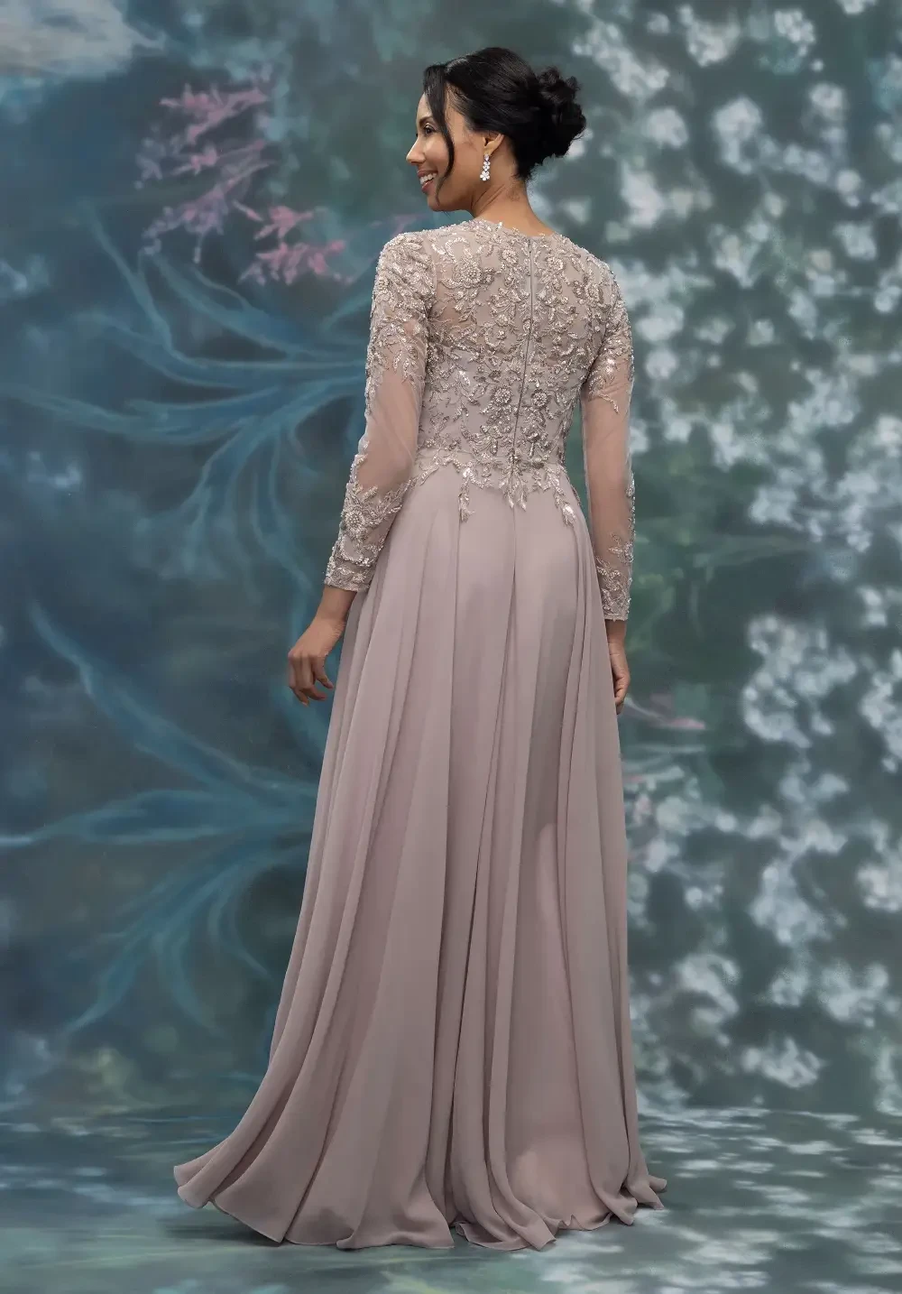 A woman wearing a long, elegant dress with intricate lace detailing on the back, standing against a soft, floral-themed background.