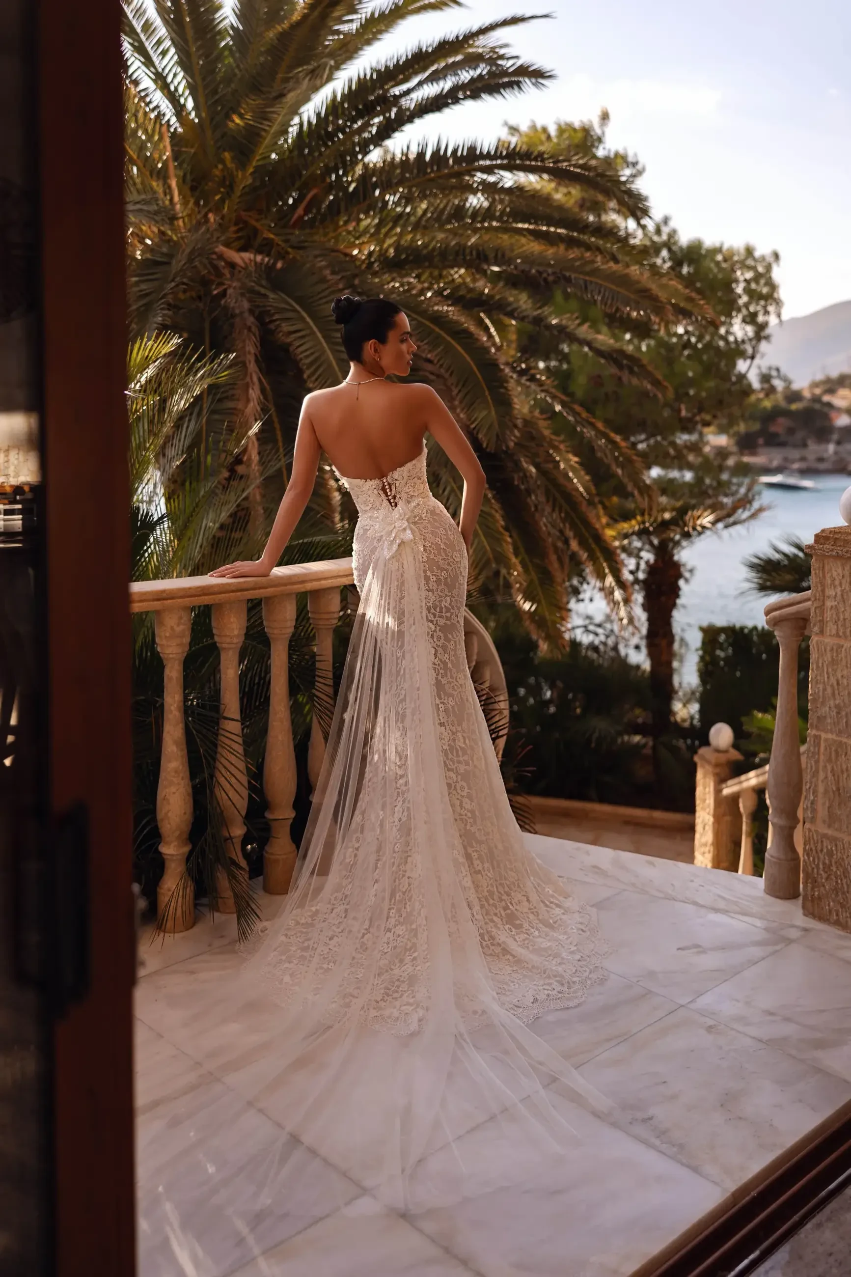 A woman in a backless wedding dress stands on a balcony overlooking palm trees and water.
