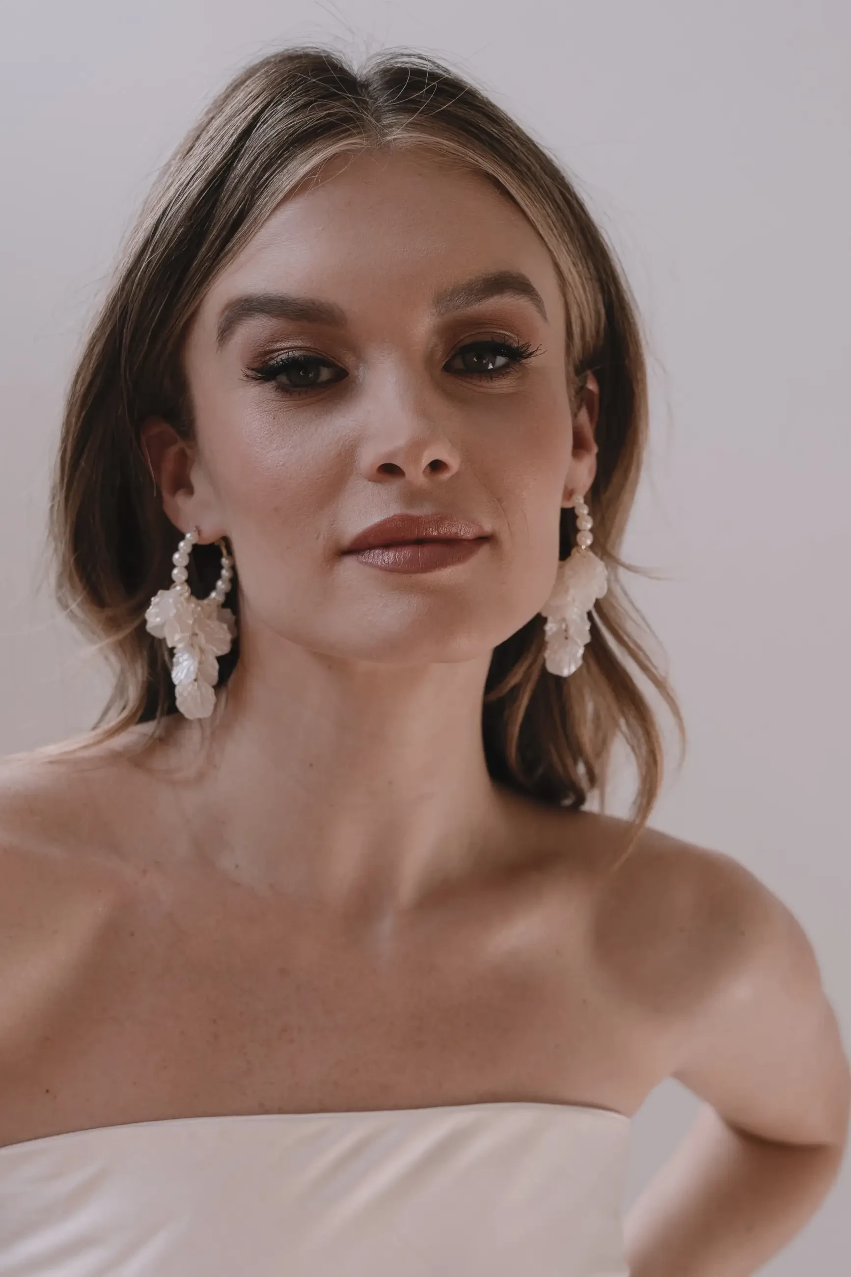 A woman with long, wavy hair poses confidently, wearing elegant pearl earrings and a strapless white top.