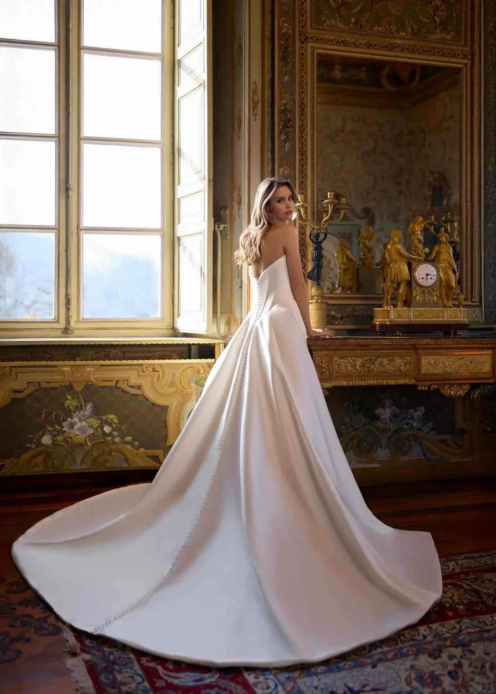 A woman in a white dress stands with her back to the camera, looking over her shoulder in a grand room with ornate decor and large windows.