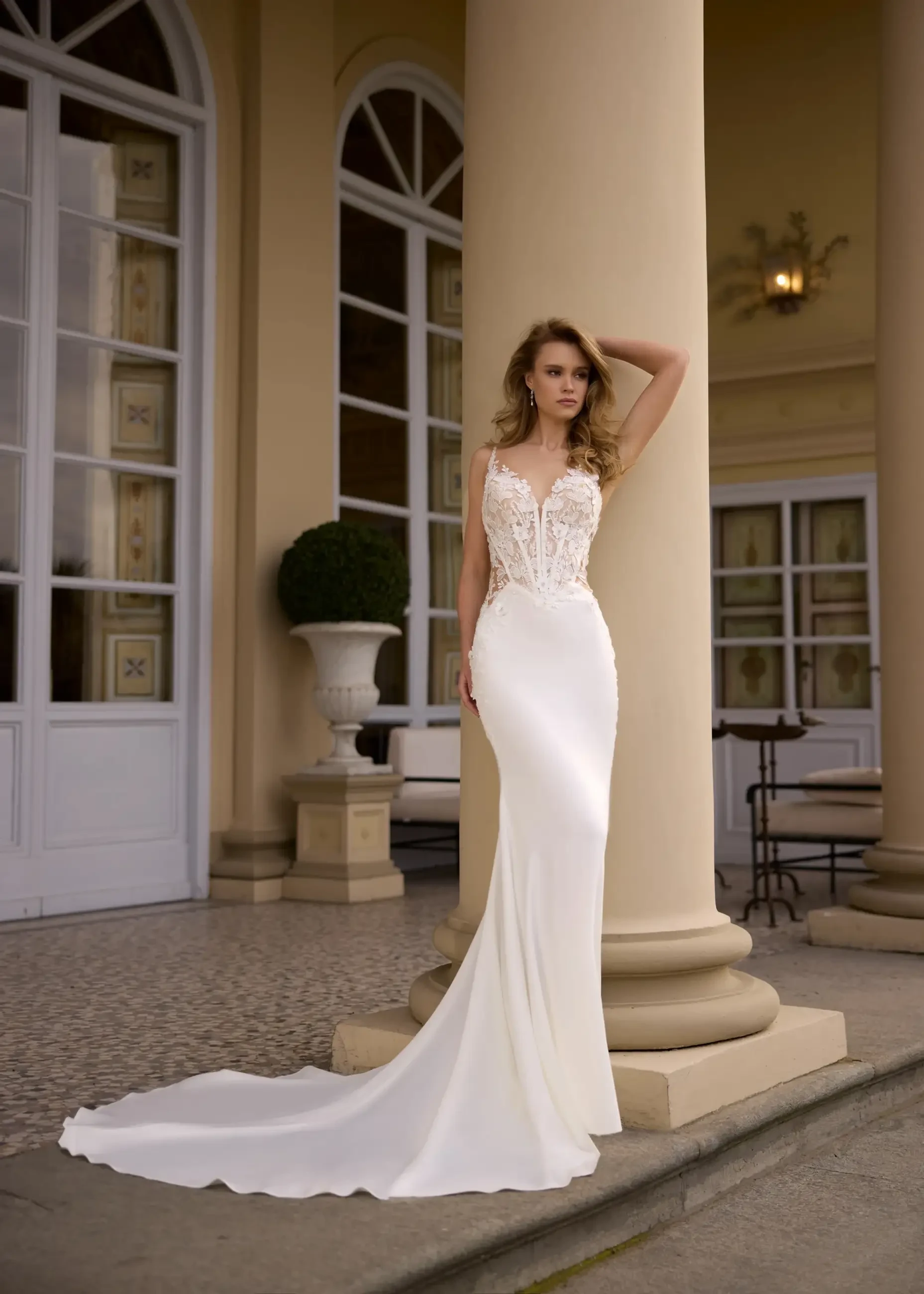 A woman stands elegantly beside a pillar, wearing a sleek white gown with intricate lace detailing on the bodice. The setting is an elegant architectural space with large windows and decorative elements.