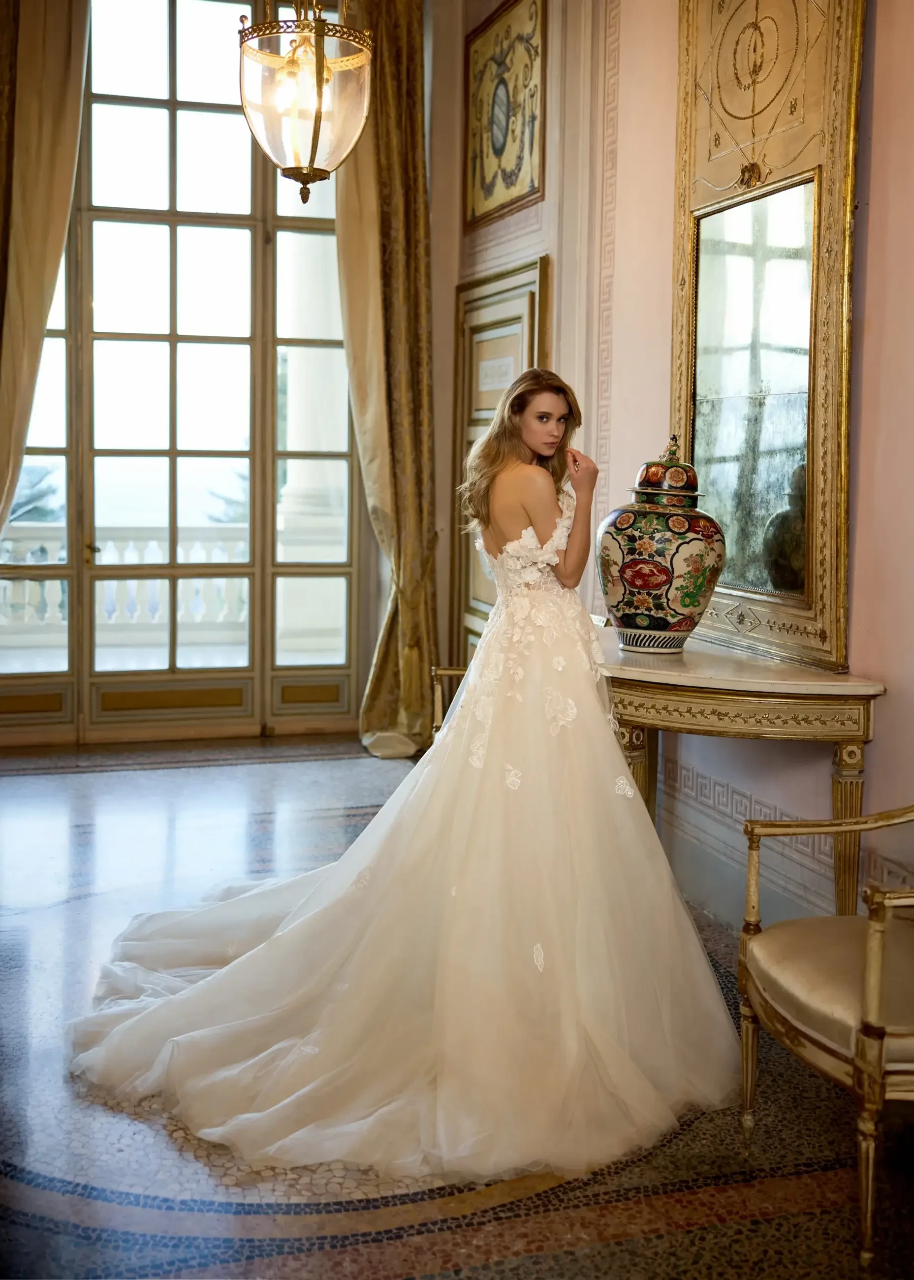 A woman in a wedding dress stands by an elegant mirror and decor in a grand room with large windows.