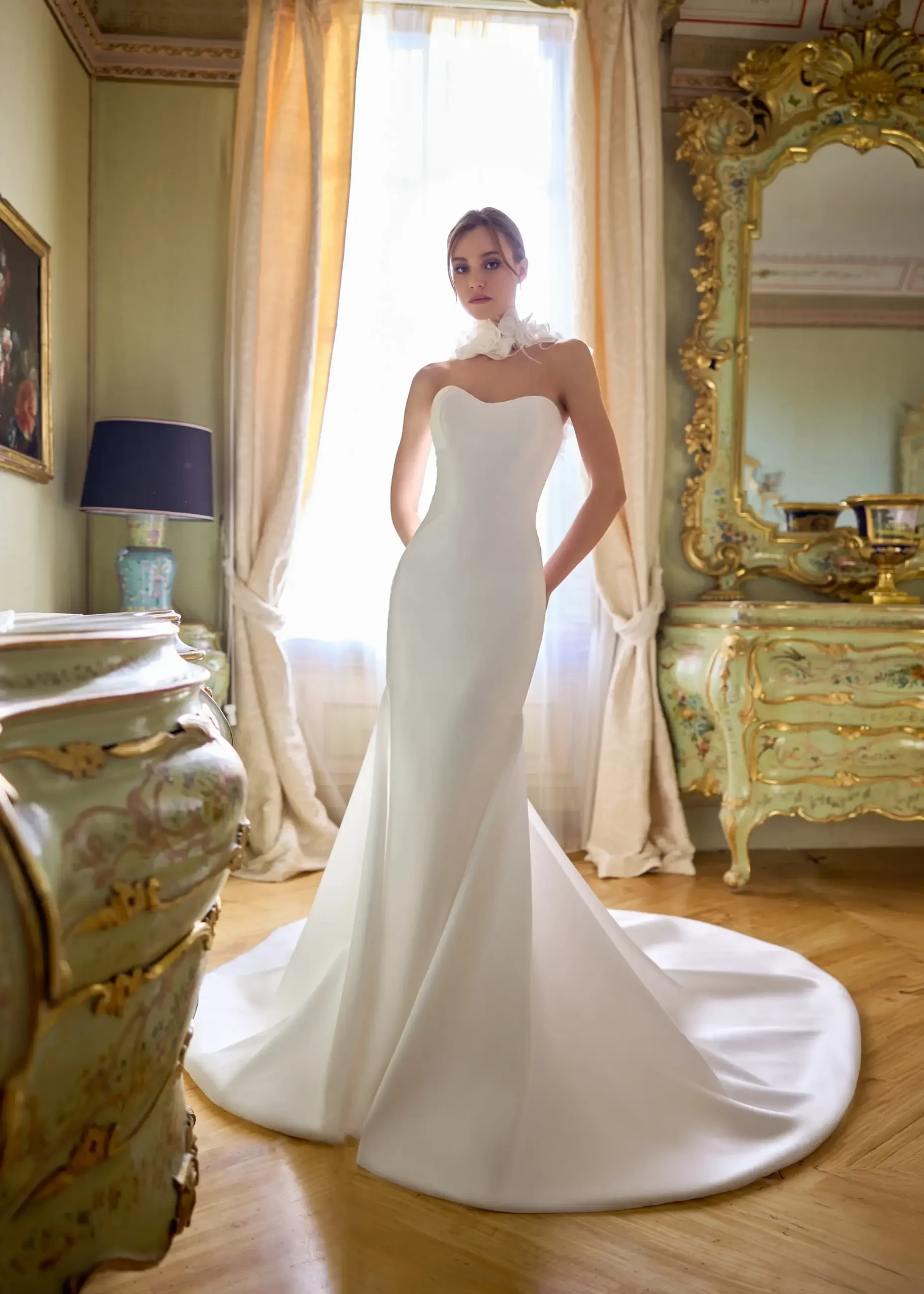 A model is posing in an elegant white wedding gown inside a beautifully decorated room with soft natural light coming through a window. The gown has a fitted silhouette and a train, and the setting includes luxurious furnishings.